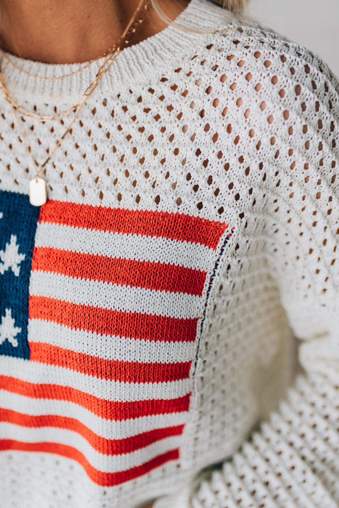 Blonde woman wearing a loose knit low gauze white sweater with drop shoulder and woven American flag at center front.