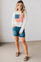 Blonde woman wearing a loose knit low gauze white sweater with drop shoulder and woven American flag at center front.