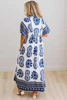Woman wearing a long, patterned dress with blue and white design against a white wall.