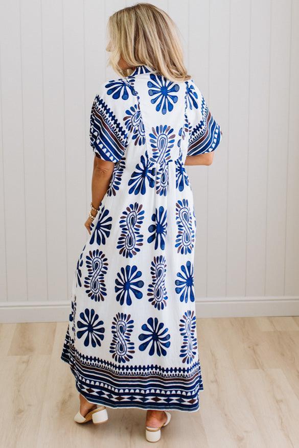 Woman wearing a long, patterned dress with blue and white design against a white wall.