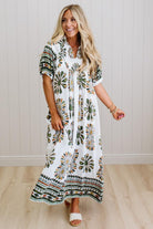 Woman wearing a long, patterned dress standing in a room with wooden flooring and a white wall.