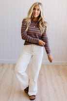 Blonde woman wearing an all over perforated mocha brown sweater with thin horizontal multi color stripes, a rolled neckline and ribbed cuff.