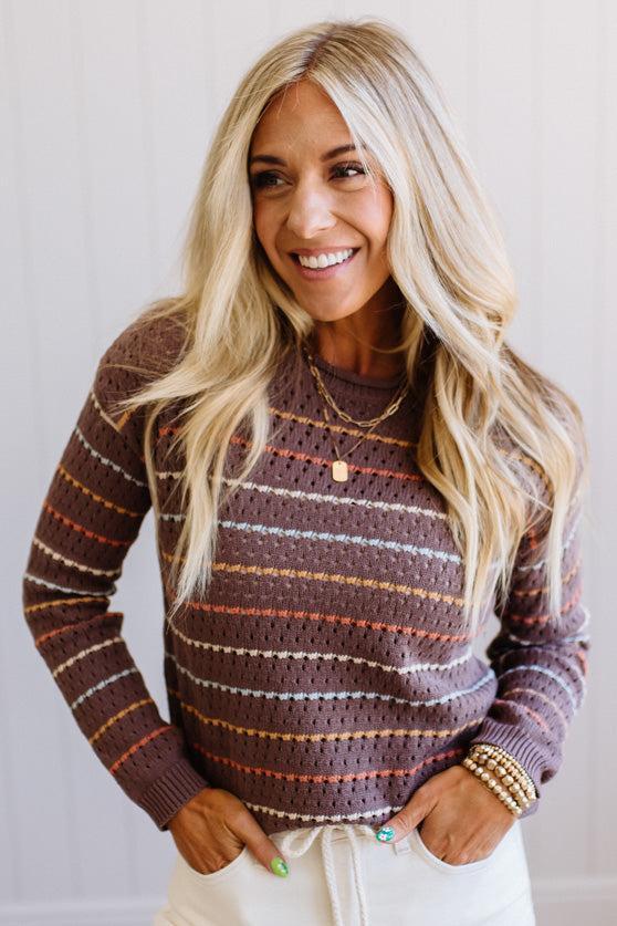 Blonde woman wearing an all over perforated mocha brown sweater with thin horizontal multi color stripes, a rolled neckline and ribbed cuff.