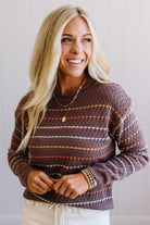 Blonde woman wearing an all over perforated mocha brown sweater with thin horizontal multi color stripes, a rolled neckline and ribbed cuff.