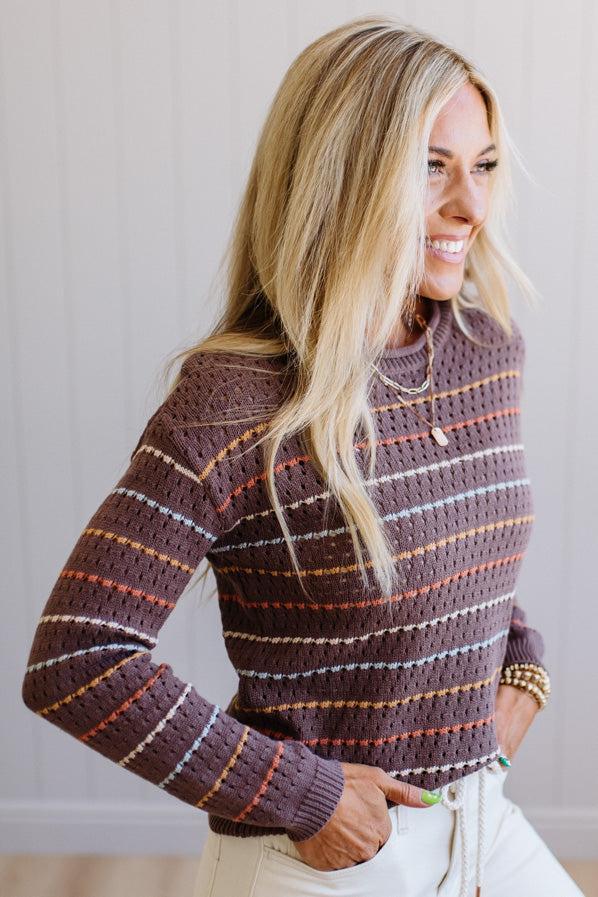 Blonde woman wearing an all over perforated mocha brown sweater with thin horizontal multi color stripes, a rolled neckline and ribbed cuff.