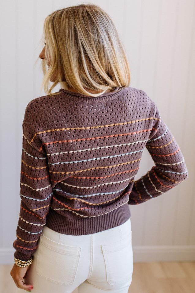 Blonde woman wearing an all over perforated mocha brown sweater with thin horizontal multi color stripes, a rolled neckline and ribbed cuff.
