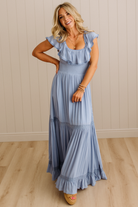 Woman wearing a light blue dress standing in a room with wooden flooring and a beige wall.