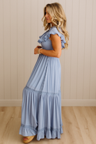 Woman wearing a light blue dress with ruffled sleeves and bottom hem against a beige wall.