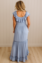 Woman wearing a light blue dress with ruffled sleeves and bottom hem against a beige wall.