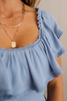 Light blue top with ruffled details and gold necklaces on a neutral background