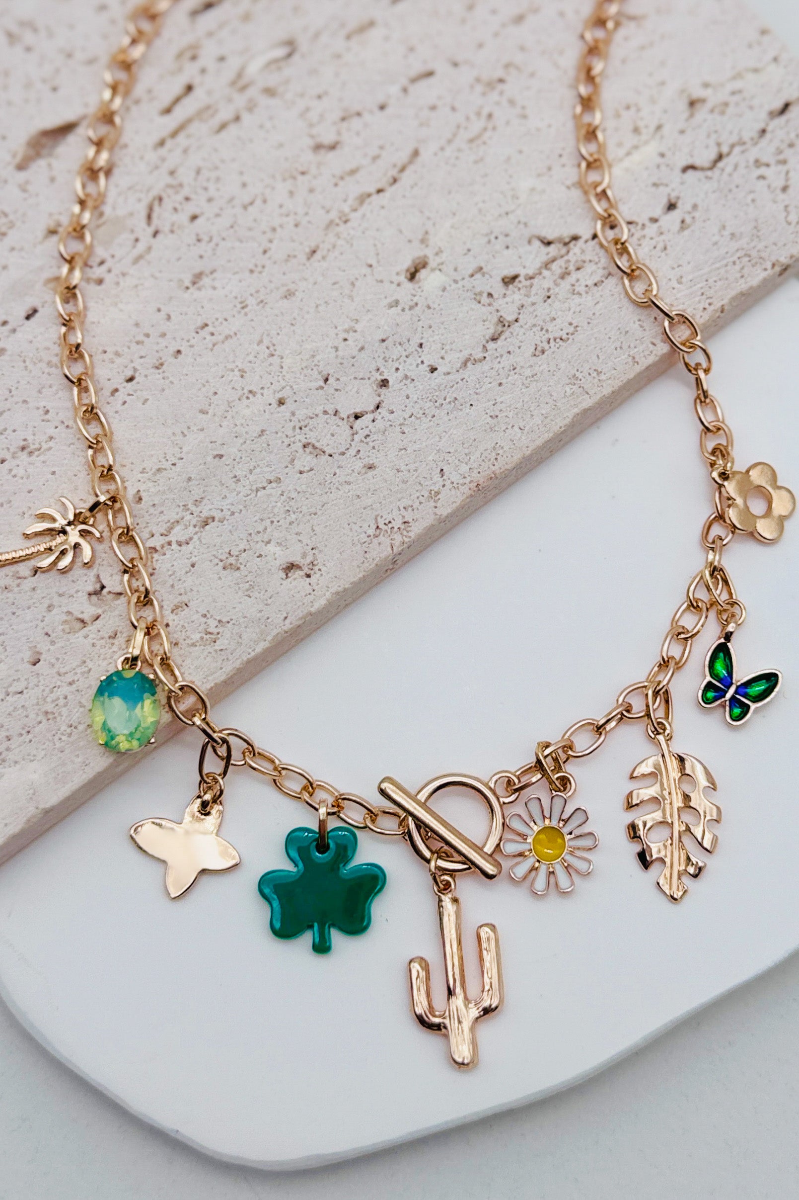 Gold necklace with various charms including a clover, cactus, and butterfly on a stone surface.