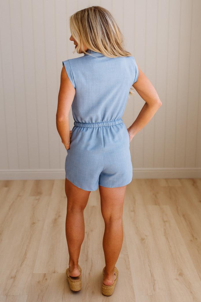 Woman wearing a light blue romper standing on a wooden floor with a neutral wall background