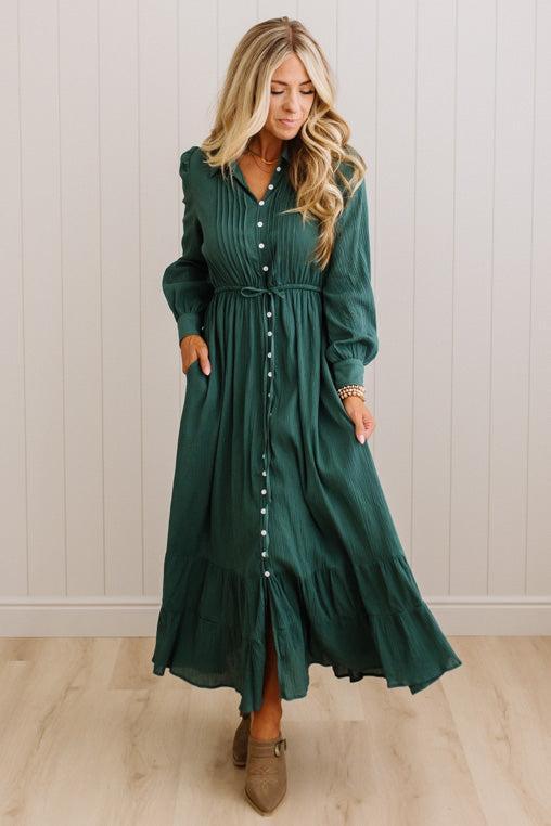 A woman wearing a long-sleeved, dark green midi dress with a button-down front and ruffled hem, standing in front of a white paneled wall and light wood floor.