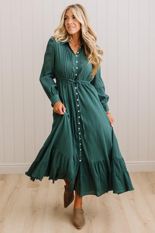 A woman modeling a long-sleeve, dark green midi dress with a button-down front, pleated bodice, and flared ruffled hem, paired with brown suede ankle boots against a light wood-paneled wall.