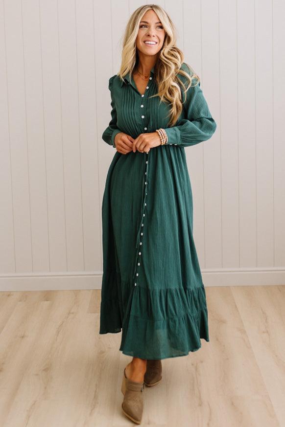 A woman wearing a dark green long-sleeve button-down maxi dress with a crinkle texture, standing in a room with light wood floors and white paneled walls.