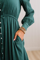 A close-up of a woman wearing a dark teal-green crinkle-textured long-sleeve dress with a button-down front and side pockets, accessorized with stacked gold beaded bracelets.