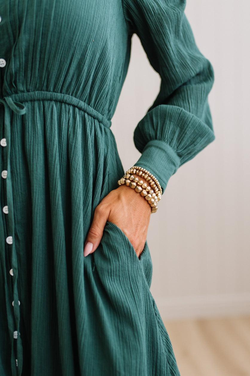 A close-up of a woman wearing a dark teal-green crinkle-textured long-sleeve dress with a button-down front and side pockets, accessorized with stacked gold beaded bracelets.