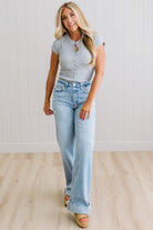 Woman wearing a gray t-shirt and light blue jeans standing on a wooden floor with a white wall background.