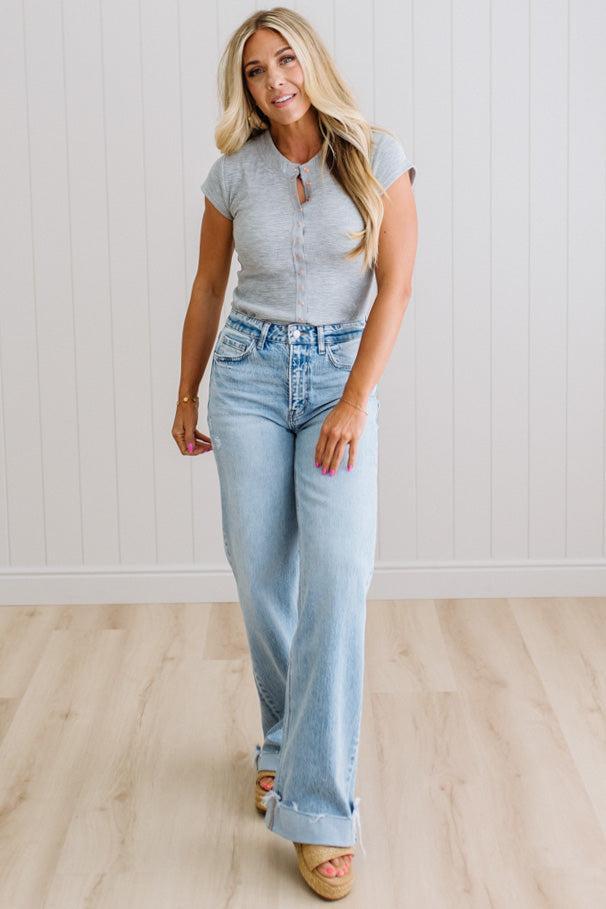 Woman wearing a gray t-shirt and light blue jeans standing on a wooden floor with a white wall background.