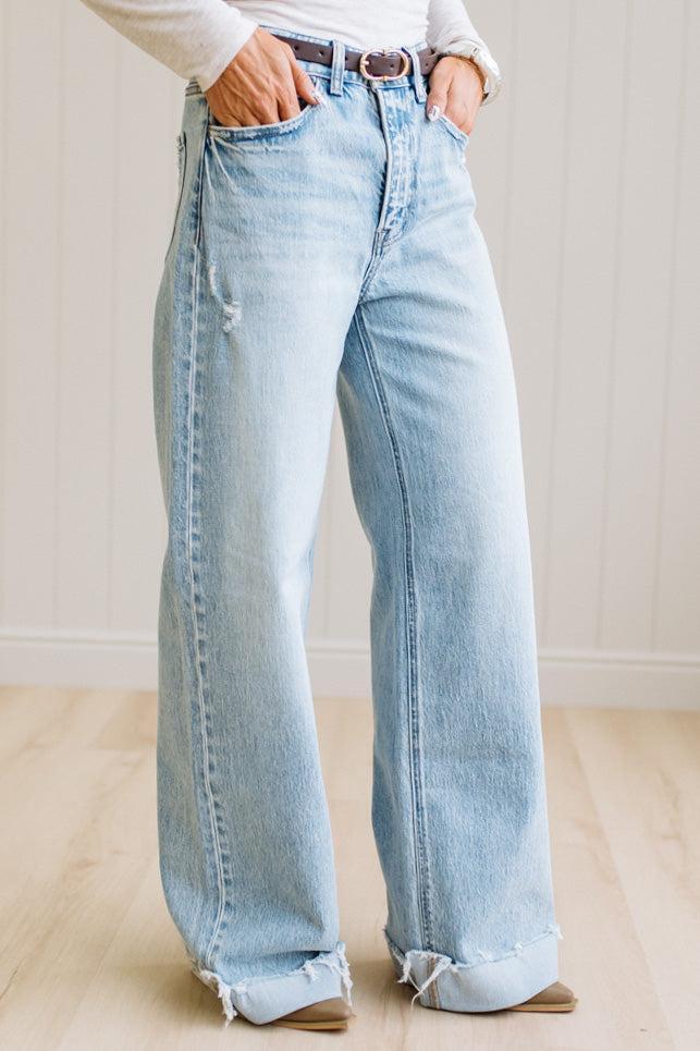 Woman wearing a gray t-shirt and light blue jeans standing on a wooden floor with a white wall background.