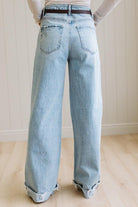 Woman wearing a gray t-shirt and light blue jeans standing on a wooden floor with a white wall background.