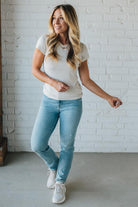 Woman wearing a white t-shirt, light blue jeans, and white sneakers against a white brick wall.