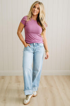 Woman wearing a purple t-shirt and light blue jeans standing against a white paneled wall.