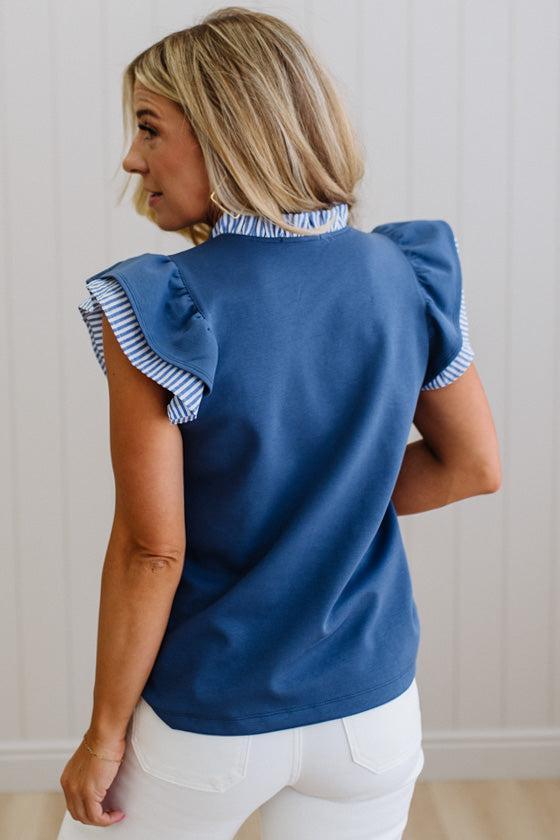 Woman wearing a blue top with ruffled sleeves and white pants against a neutral background