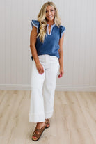 Woman wearing a blue top and white pants standing against a light wooden paneled wall.