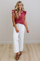 Woman wearing a pink blouse with ruffled sleeves and white pants against a light wooden panel background.