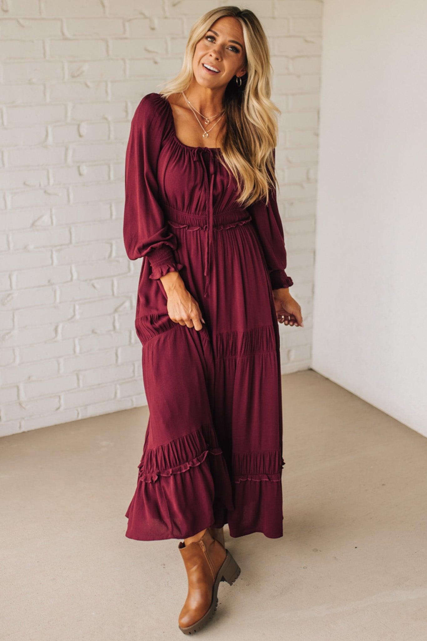 Woman wearing a burgundy dress with long sleeves and ruffled hem against a white brick wall.
