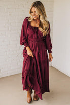 Woman wearing a burgundy dress with long sleeves and a white brick wall background