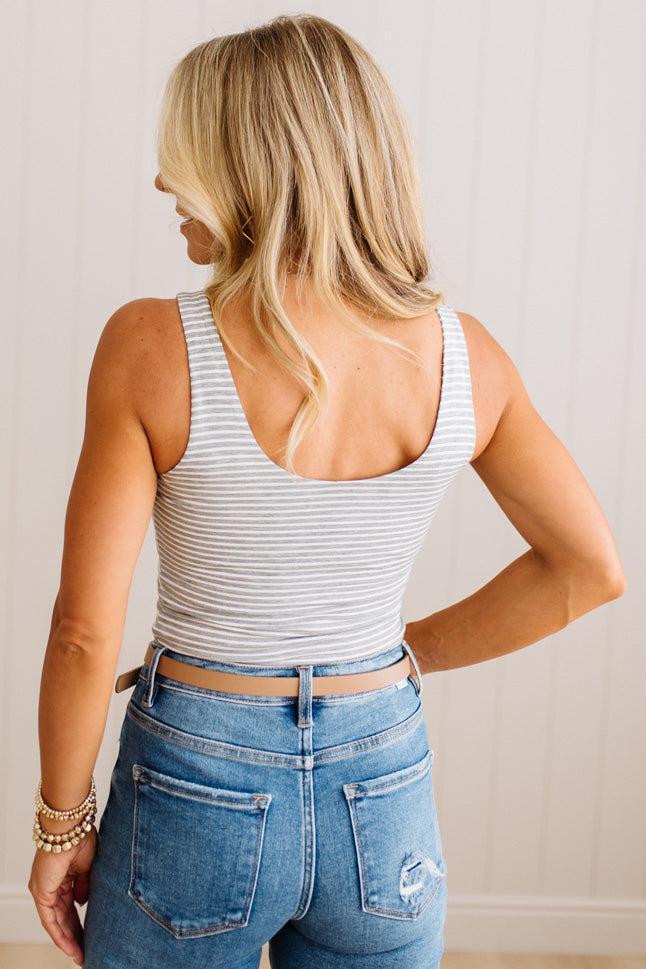 A woman shown from the back wearing a white and gray striped ribbed tank top and blue jeans, standing against a light beige paneled wall.