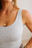 A close-up of a woman wearing layered gold necklaces, including a rectangular dog tag pendant, over a gray and white striped tank top.