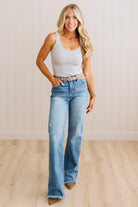 A woman modeling a sleeveless white and gray striped tank top with light-wash blue flare jeans and a tan belt, standing in a bright room with light wood flooring and white paneled walls.