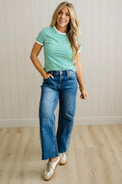 Woman wearing a green and white striped shirt and blue jeans standing against a light wooden paneled wall.
