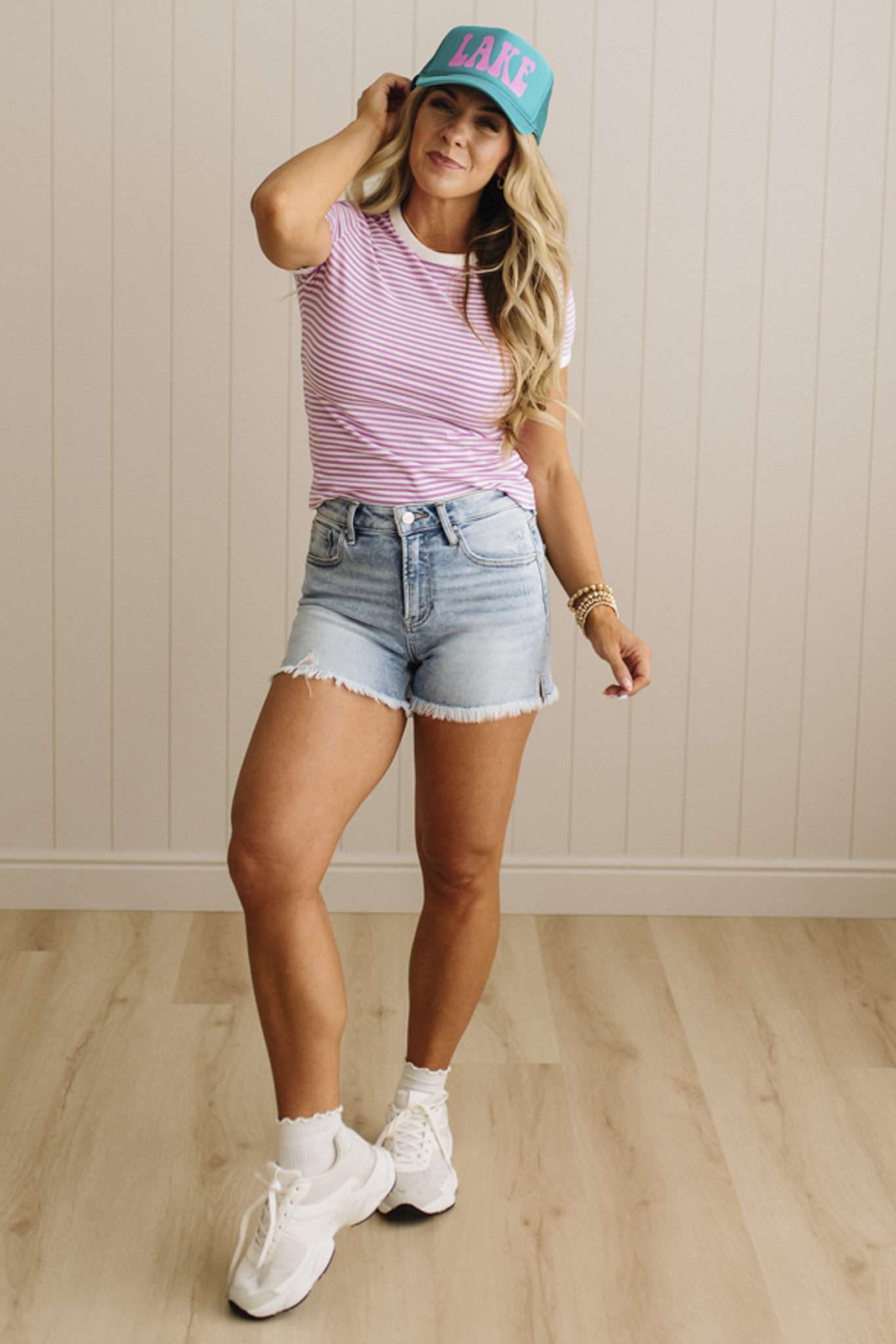 Woman wearing a striped shirt, denim shorts, and sneakers with a teal cap indoors.