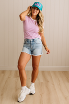 Woman wearing a striped shirt, denim shorts, and sneakers with a teal cap indoors.
