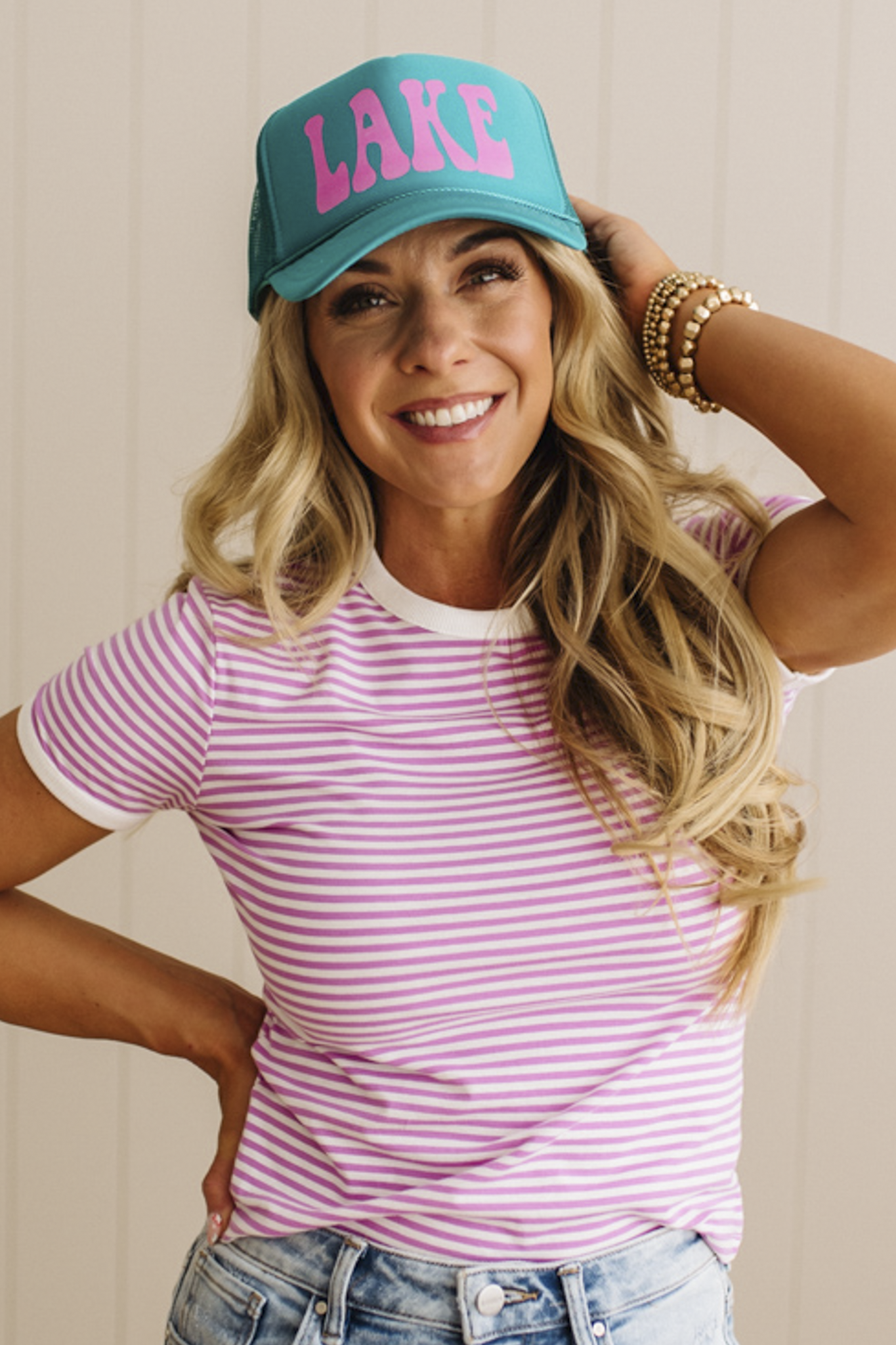 Woman wearing a turquoise cap with 'LAKE' in pink and a striped shirt against a neutral background