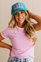 Woman wearing a turquoise cap with 'LAKE' in pink and a striped shirt against a neutral background