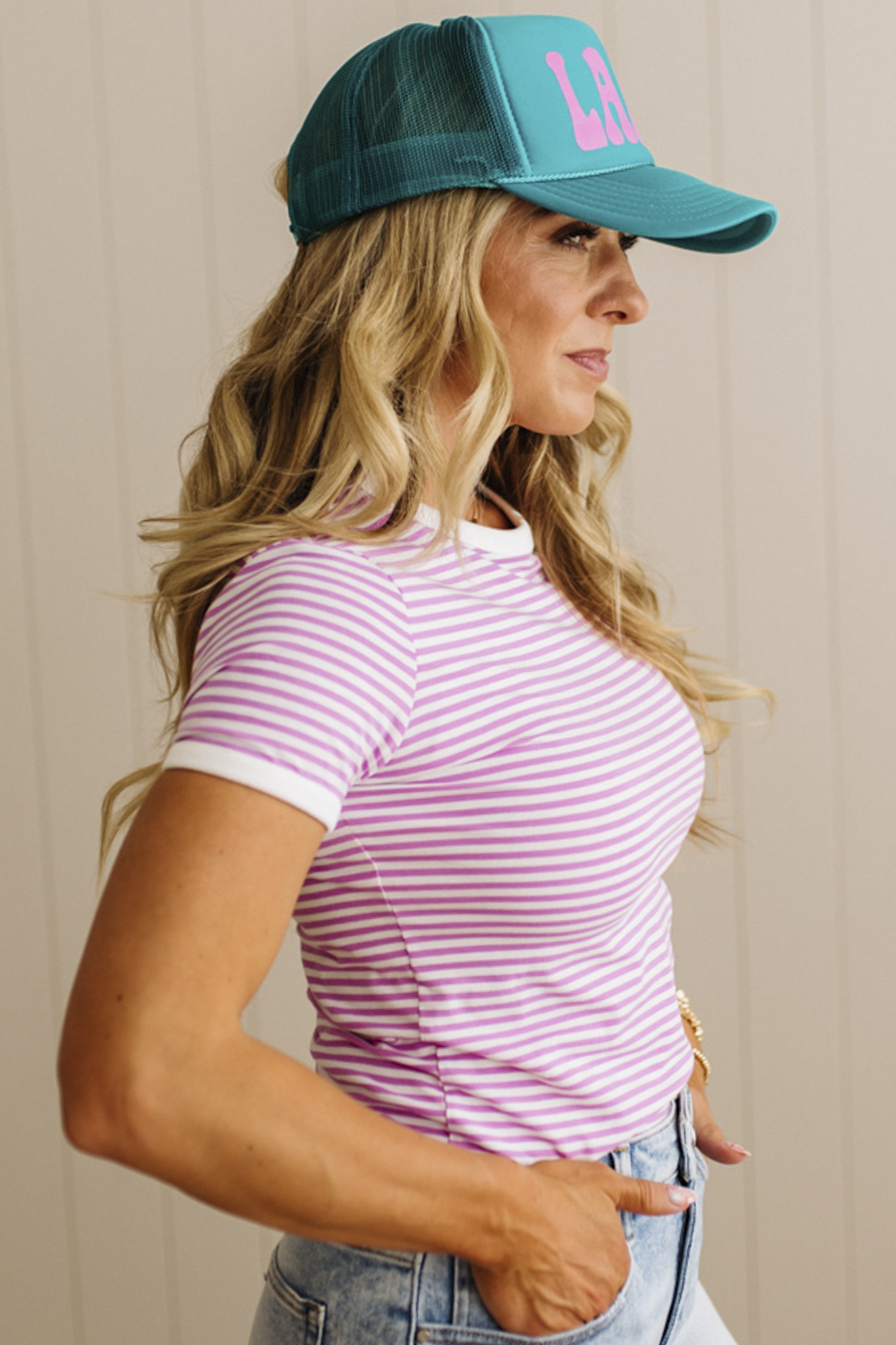 Woman wearing a pink striped shirt and teal cap with pink logo against a beige background