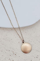 A gold chain necklace with a flat, circular disc pendant, displayed on a textured beige surface with a white ceramic plate in the background.