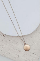 A delicate rose gold chain necklace with a small, textured circular disc pendant, displayed on a white ceramic plate over a speckled beige stone surface.