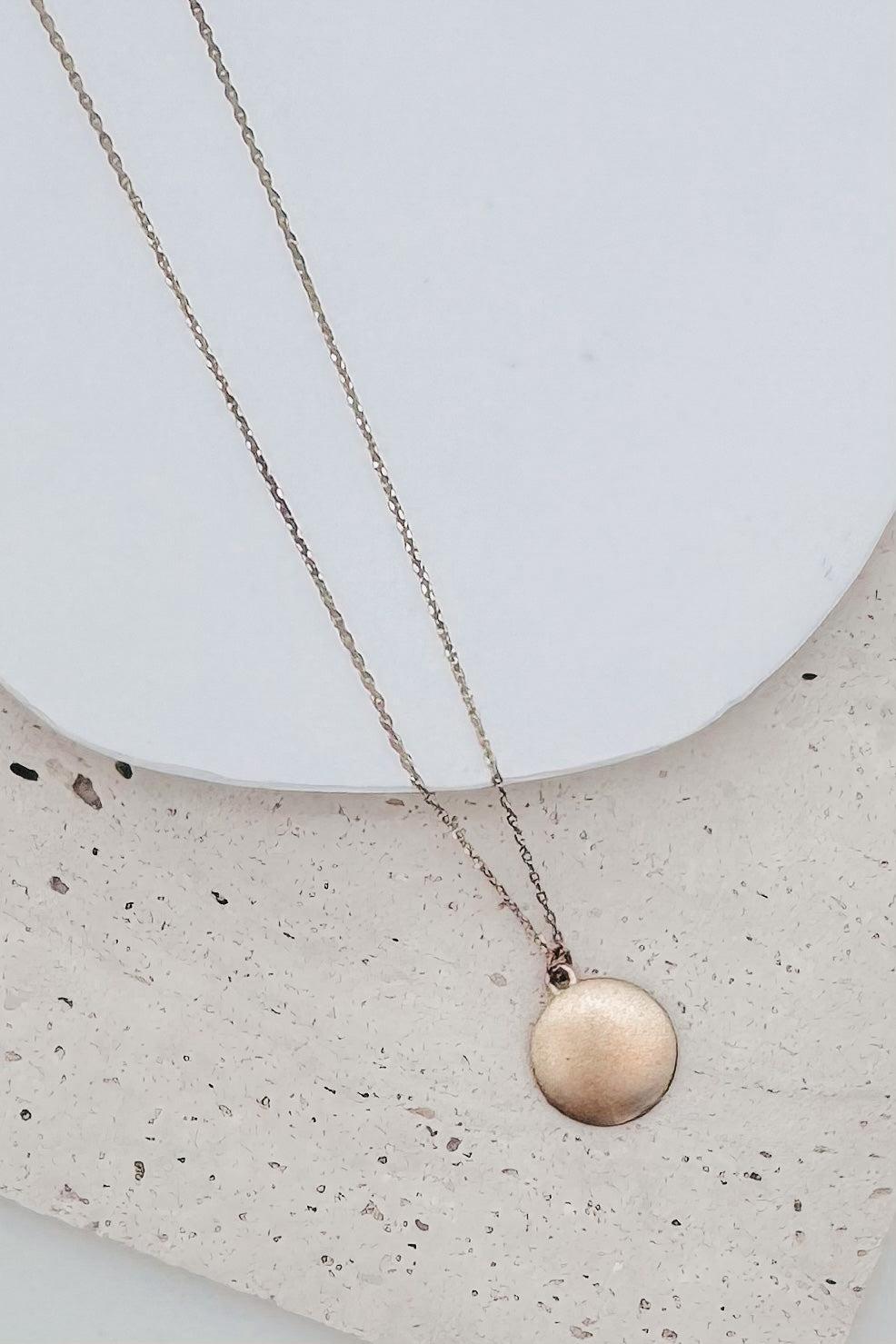 A delicate rose gold chain necklace with a small, textured circular disc pendant, displayed on a white ceramic plate over a speckled beige stone surface.