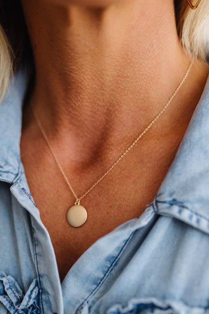 A close-up of a person's neck and chest wearing a delicate gold chain necklace with a small, flat circular disc pendant. The person is wearing a light blue denim shirt.