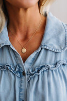 A close-up of a woman wearing a delicate gold chain necklace with a small, flat circular disc pendant, styled with a light blue denim shirt.