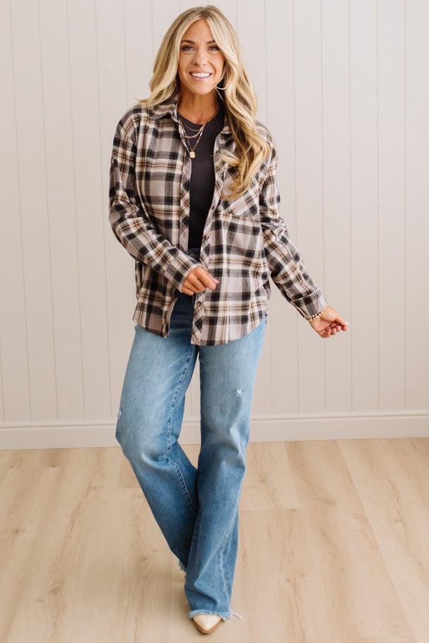 A woman modeling a brown and beige plaid button-down flannel shirt over a black top and blue jeans, standing in a room with light wood floors and white paneled walls.