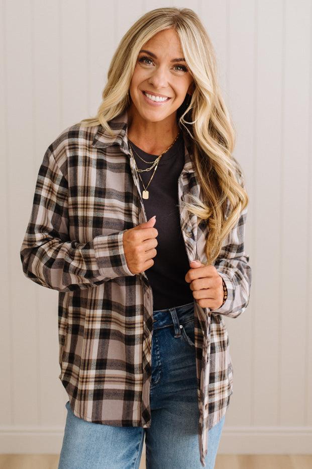 A smiling blonde woman wearing a brown, black, and beige plaid button-up shirt over a black t-shirt and blue jeans, standing against a light beige paneled wall.