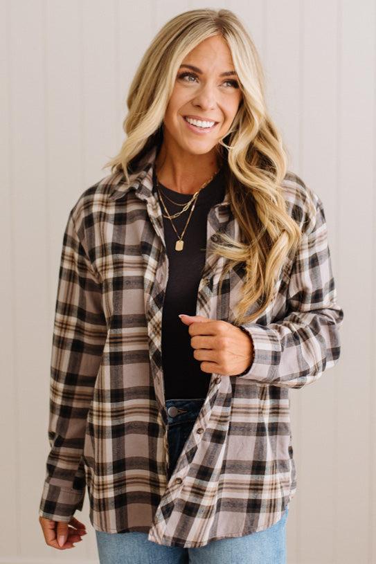 A woman wearing a long-sleeve plaid button-up shirt in brown, beige, and black over a black top and blue jeans, standing against a light beige paneled wall.
