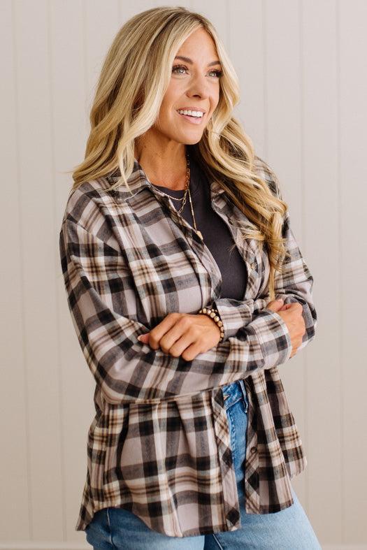 A woman wearing a brown, beige, and black plaid button-up shirt over a dark top and blue jeans, standing against a light beige paneled wall.